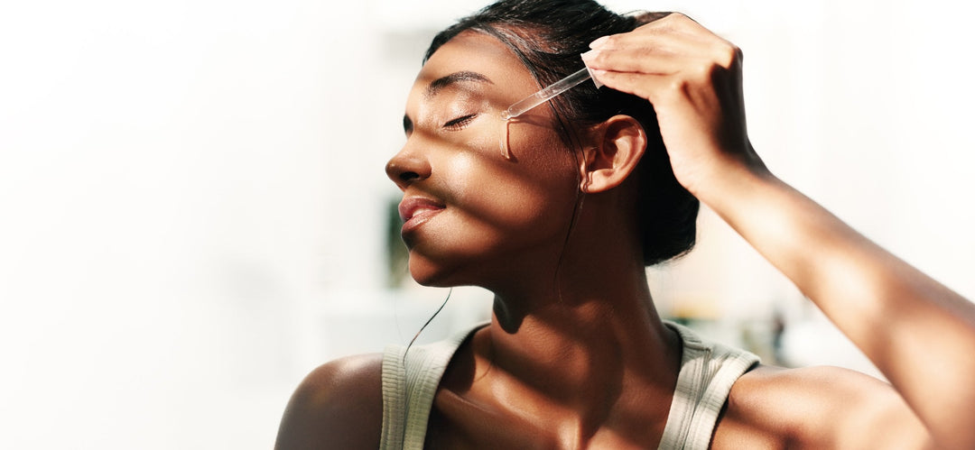 A woman applies vitamin C serum to her cheek with a glass dropper, in bright natural daylight.